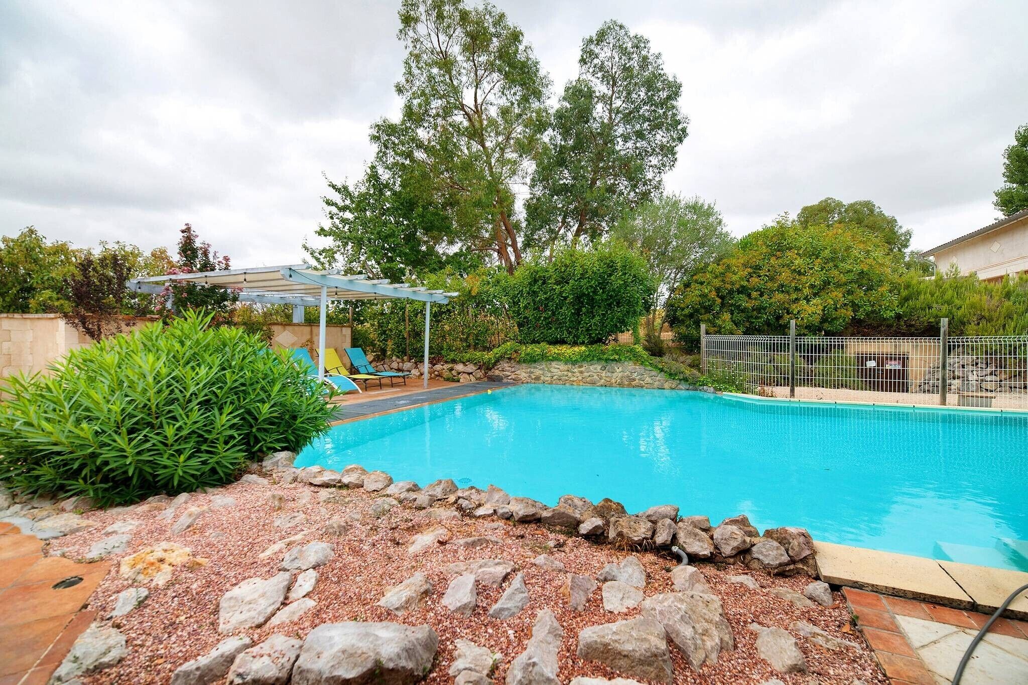 A swimming pool with a stone border, surrounded by greenery and a pergola.