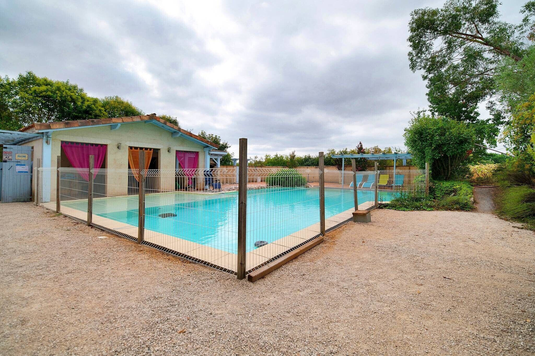 Una piscina con área techada, un edificio con una puerta que dice 'PRESCINE' y un sendero de grava.