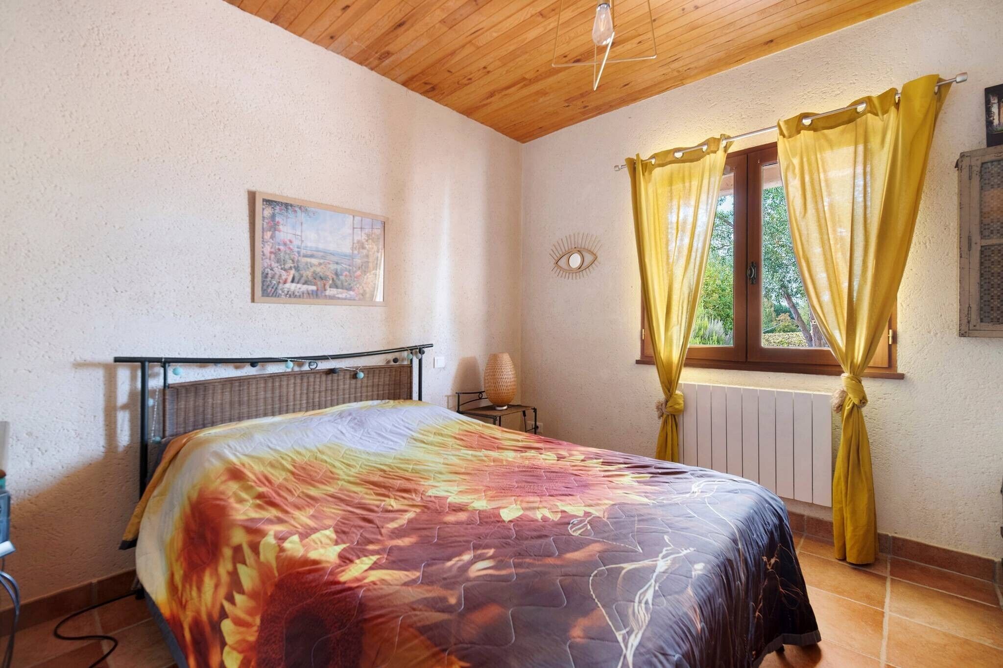 Een slaapkamer met een bed, een radiator, een raam met gordijnen, een foto aan de muur en een plafondventilator.