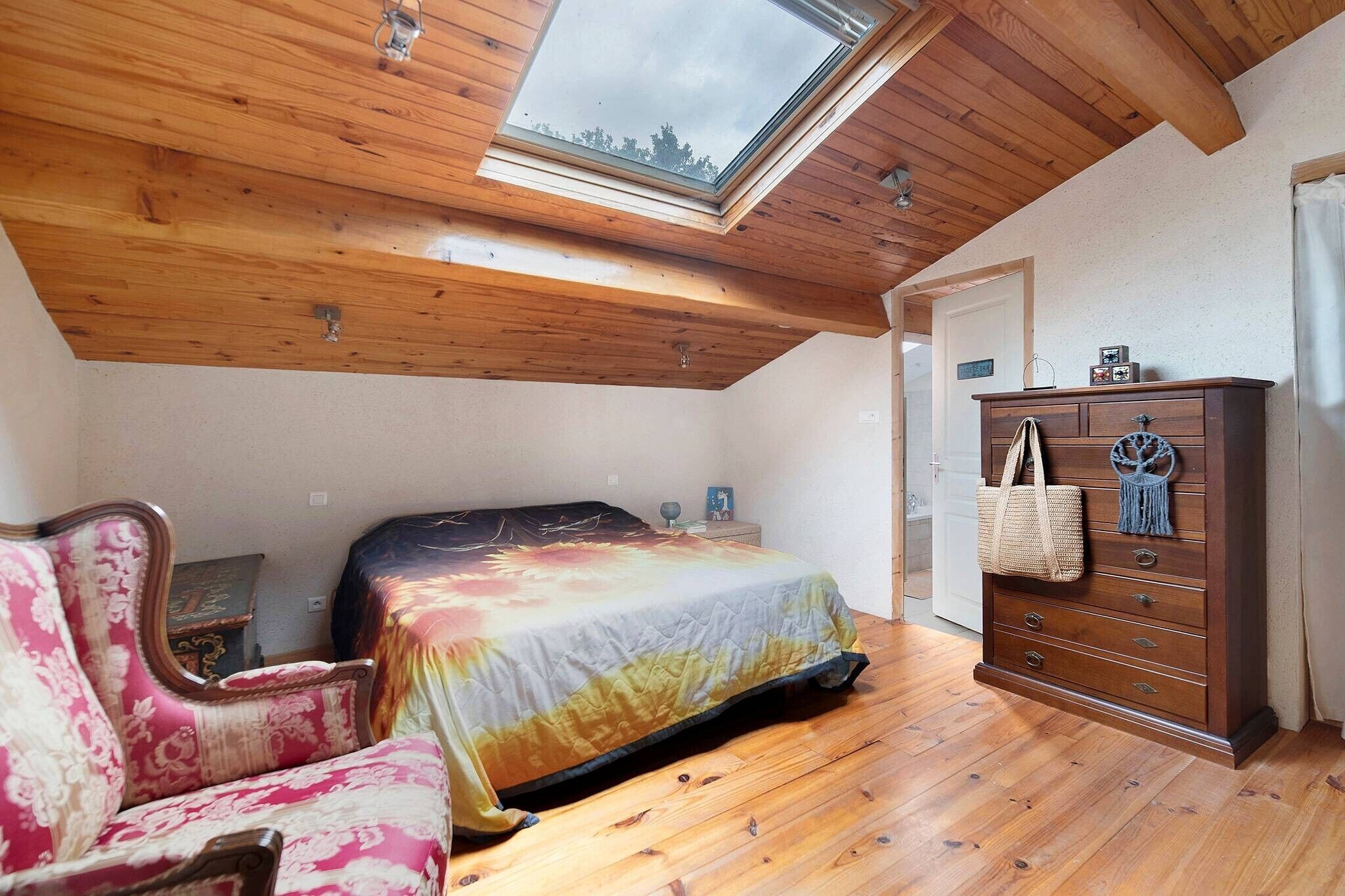 A bedroom with a skylight, wooden floor, a bed with a colorful bedspread, a wooden dresser, and a chair.