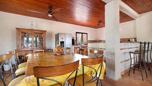 Un coin repas avec une table et des chaises en bois, un ventilateur de plafond, et un espace cuisine équipé d’un réfrigérateur et d’un évier.
