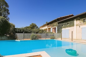 A swimming pool with a clear blue water, surrounded by a fence, and a building with a tiled roof and a garden.