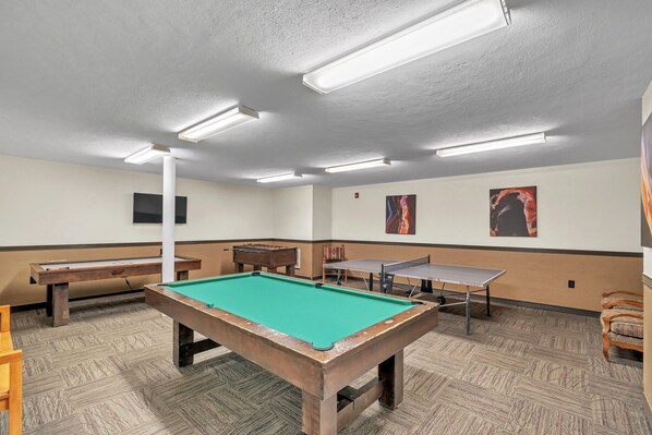 Una mesa de billar, una mesa de ping pong y una mesa de fútbolín en una sala con alfombra estampada y paredes blancas.