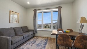 A room with a grey sofa, a dining table with chairs, a window with curtains, and a lamp.