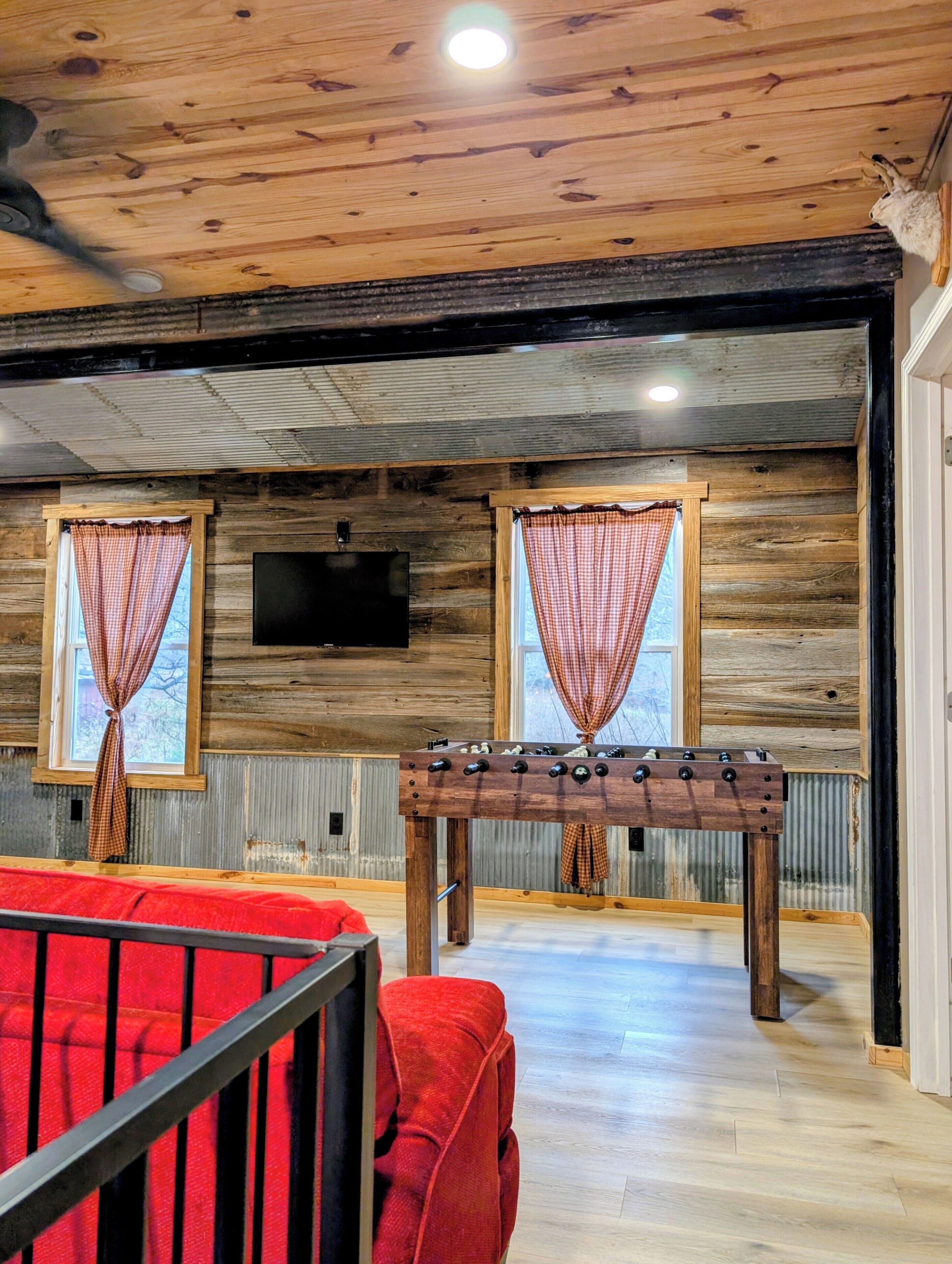 A cozy room with wooden walls and a ceiling, featuring a foosball table, two windows with checkered curtains, and a flat-screen TV mounted on the wall.