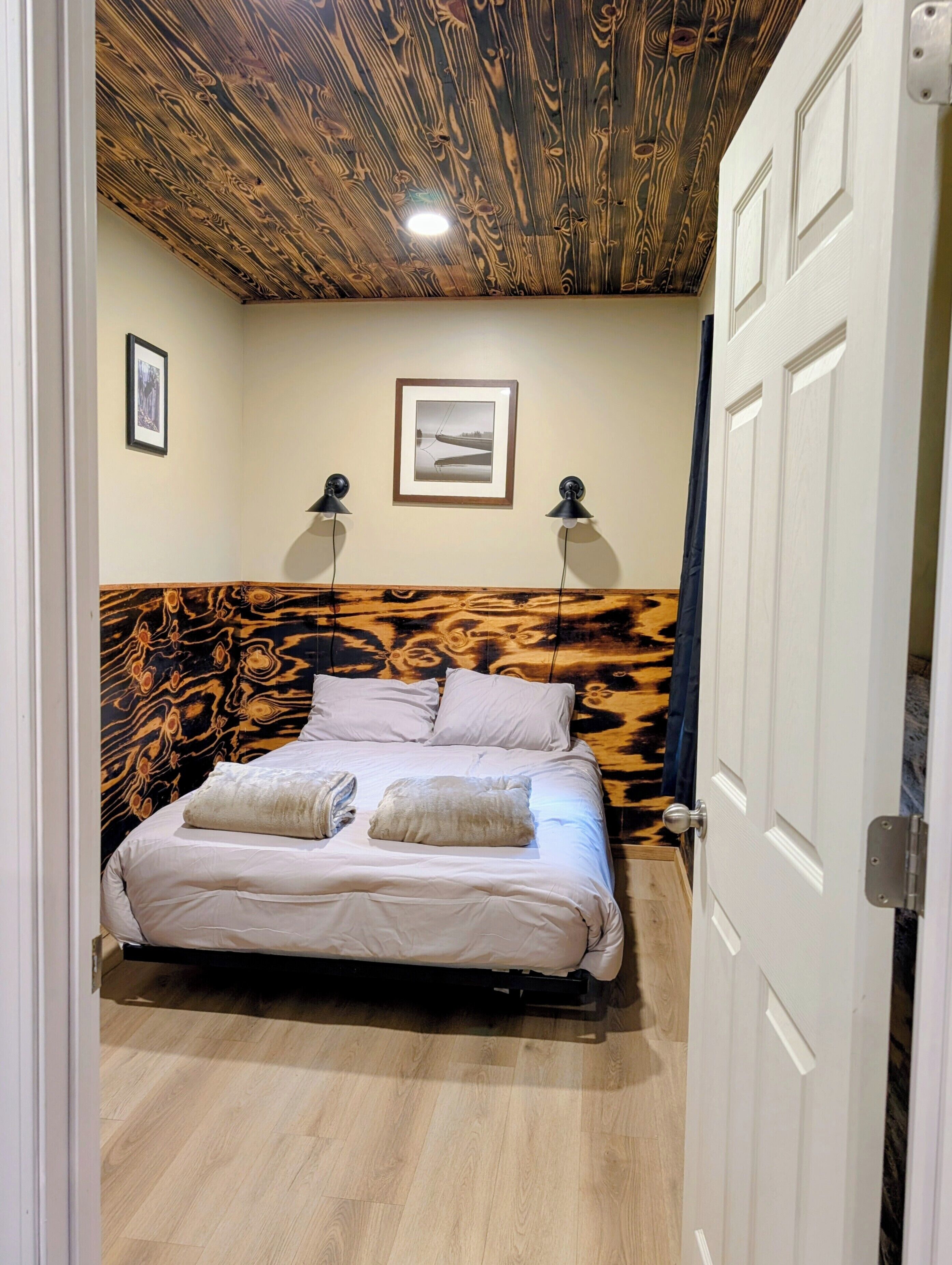 A bedroom with a bed, two bedside lamps, a framed picture on the wall, and a wooden ceiling.