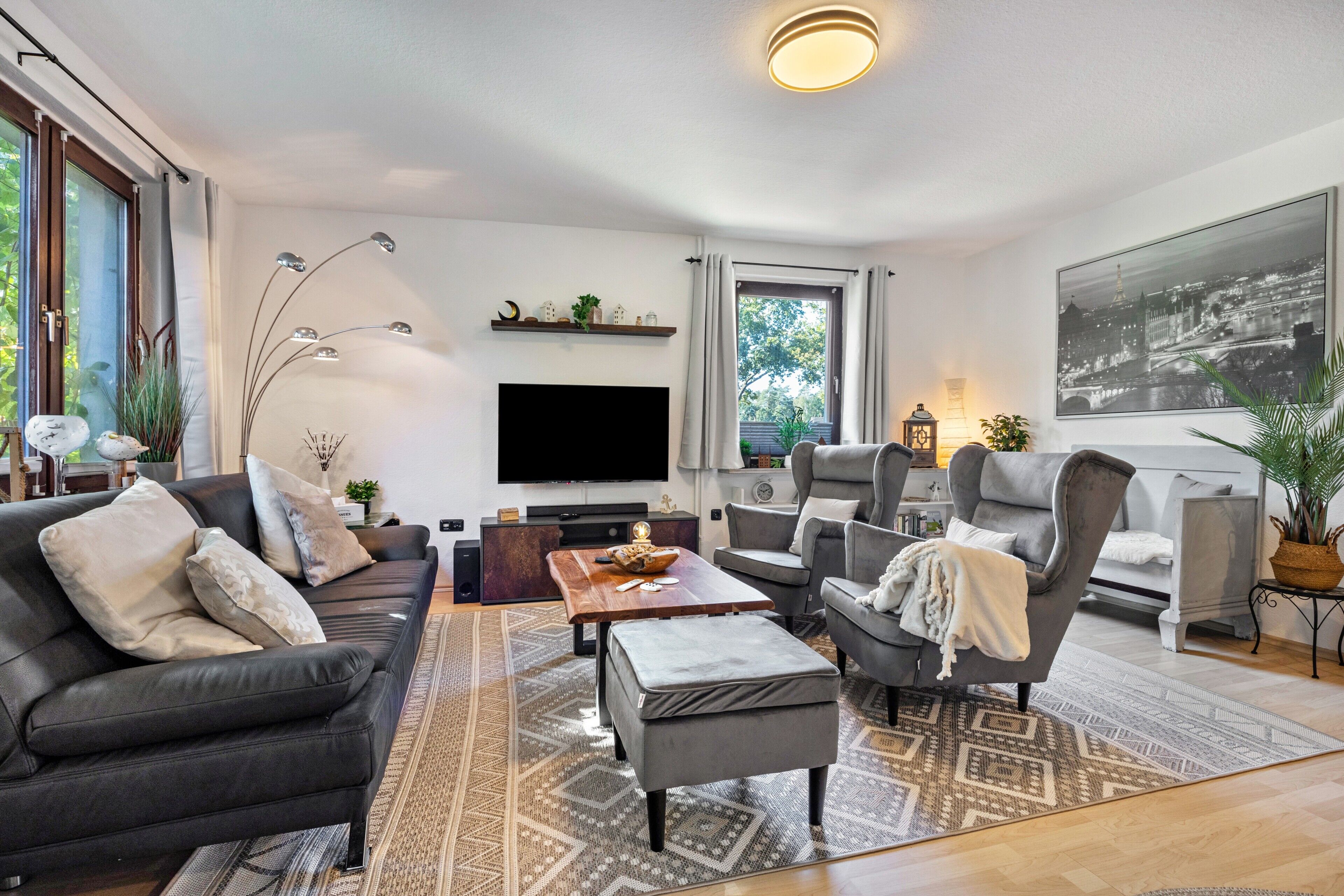 Een moderne woonkamer met een bank, fauteuils, een salontafel, een flatscreen televisie en een groot raam met gordijnen.