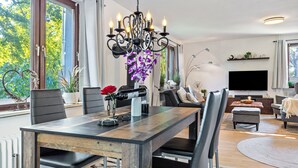 Una sala de estar moderna con una mesa de comedor, sillas, una araña, un televisor y un ventanal con vistas a árboles.