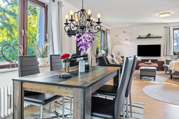 A modern living room with a dining table, chairs, a chandelier, a TV, and a large window with a view of trees.