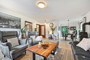 A modern living room with a wooden coffee table, two grey armchairs, a black sofa, and a dining area with a table and chairs.
