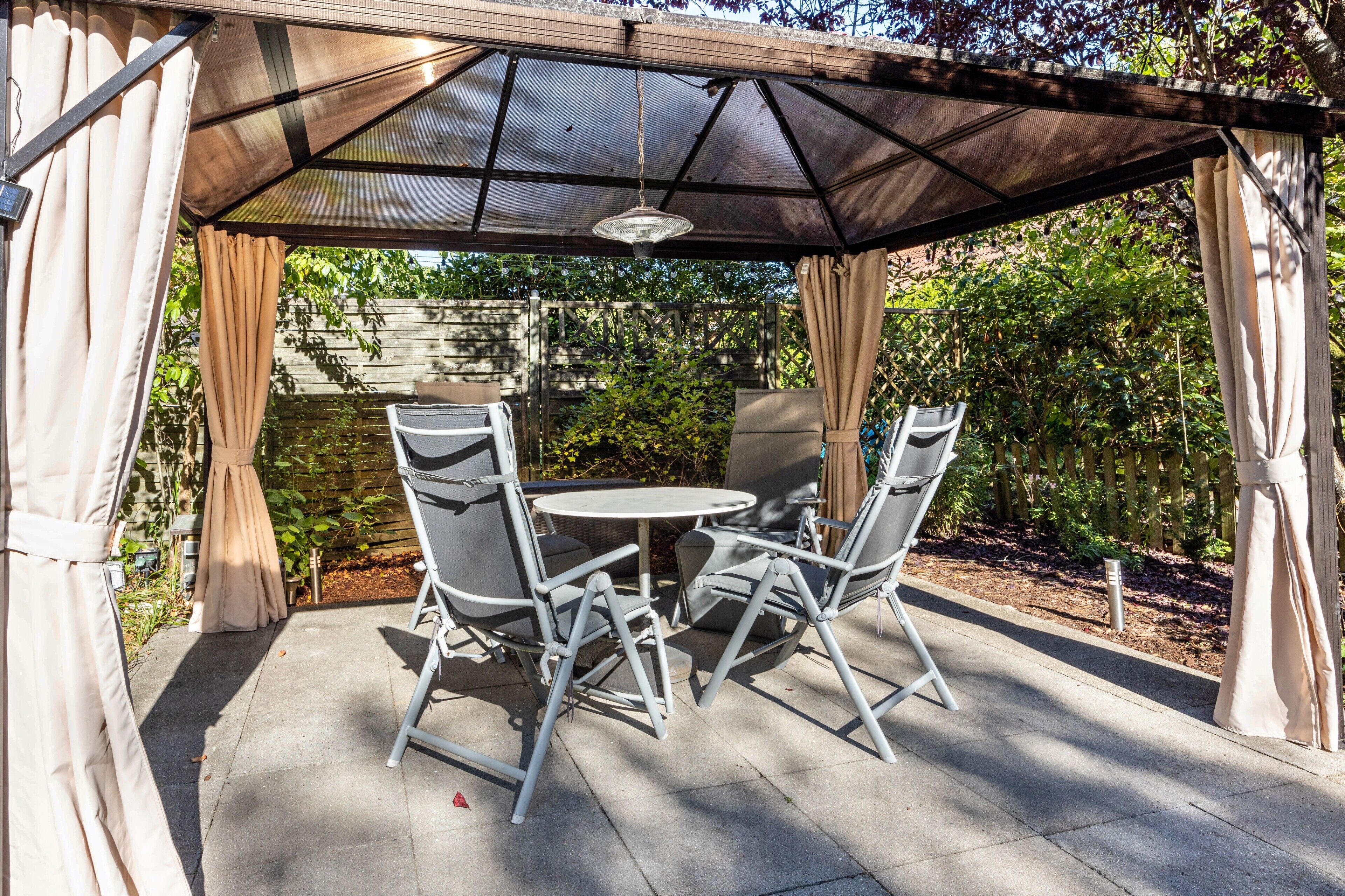 Une terrasse couverte avec une table et des chaises, un luminaire suspendu et une clôture en bois en arrière-plan.