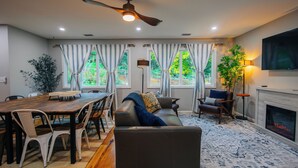 Una sala de estar moderna con comedor, ventanas grandes con cortinas, chimenea y ventilador de techo.