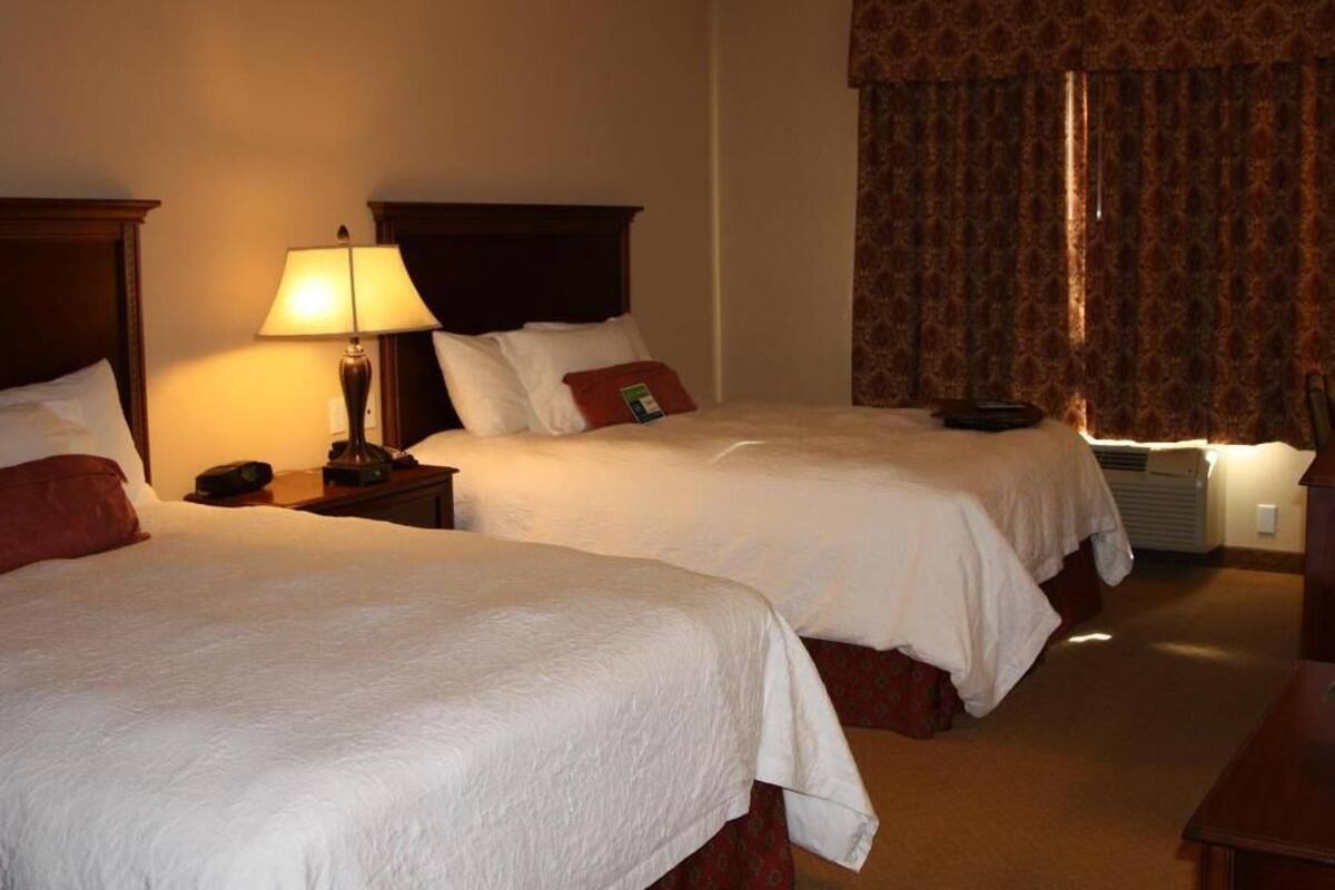 A hotel room with two beds, a bedside table with a lamp, a heater, and a window with curtains.