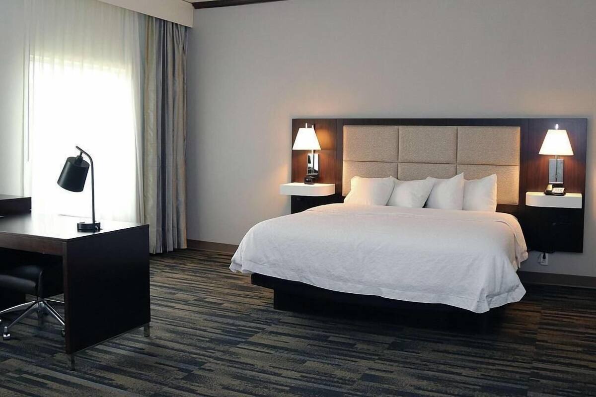 A hotel room with a bed, two bedside lamps, a desk with a chair, and a large window with curtains.