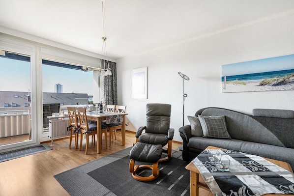A modern living room with a sofa, a dining table with chairs, a rocking chair, and a view of the beach through large windows.