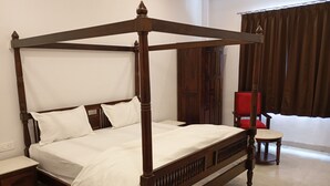 A four-poster bed with white linens, a wooden nightstand, a red chair, and a window with curtains. - Har Niwas (Udaipur)