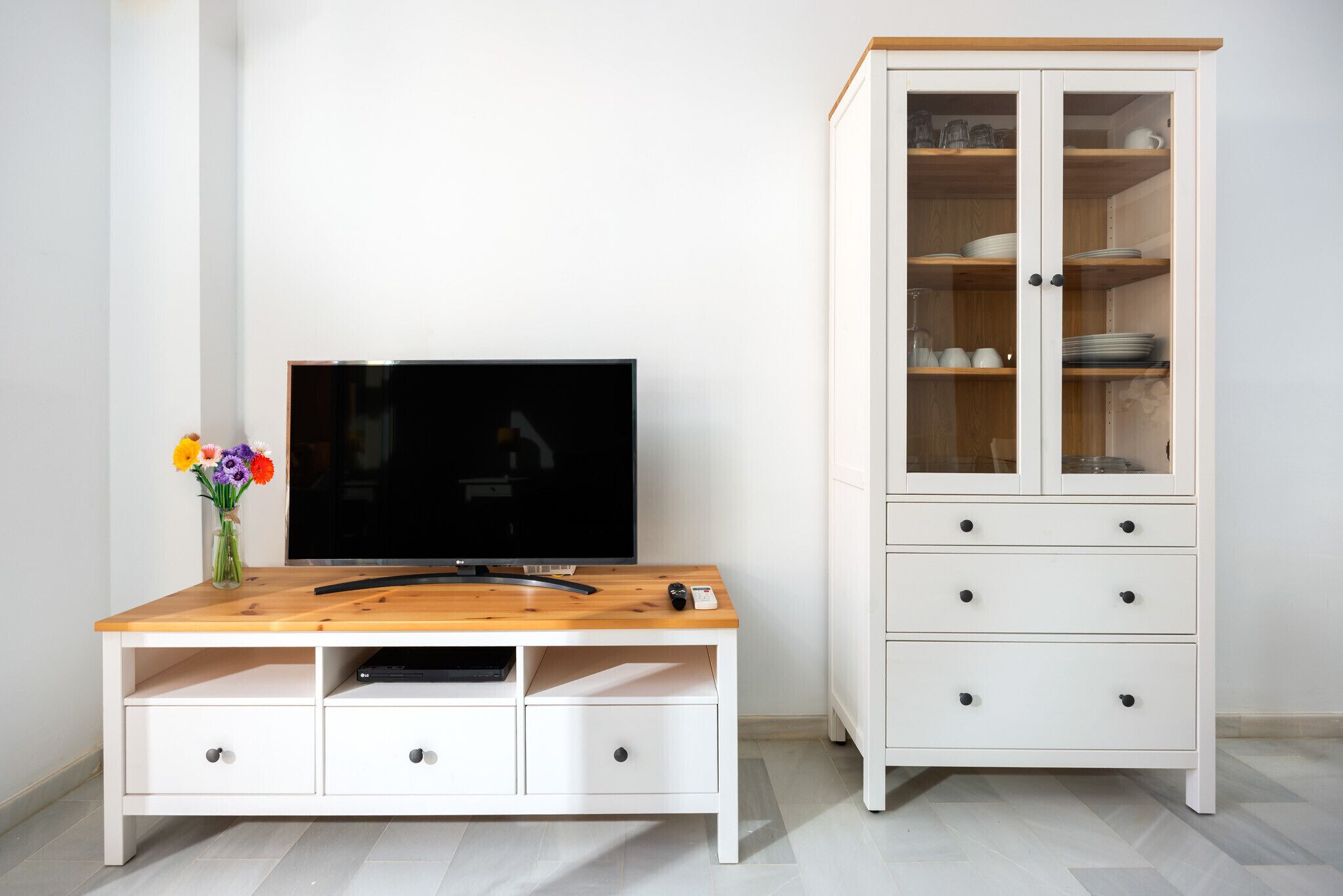 Une pièce comprenant un meuble de rangement en bois abritant un téléviseur à écran plat, une télécommande et un lecteur DVD. À côté se trouve un meuble blanc aux portes vitrées, contenant de la vaisselle et des verres.