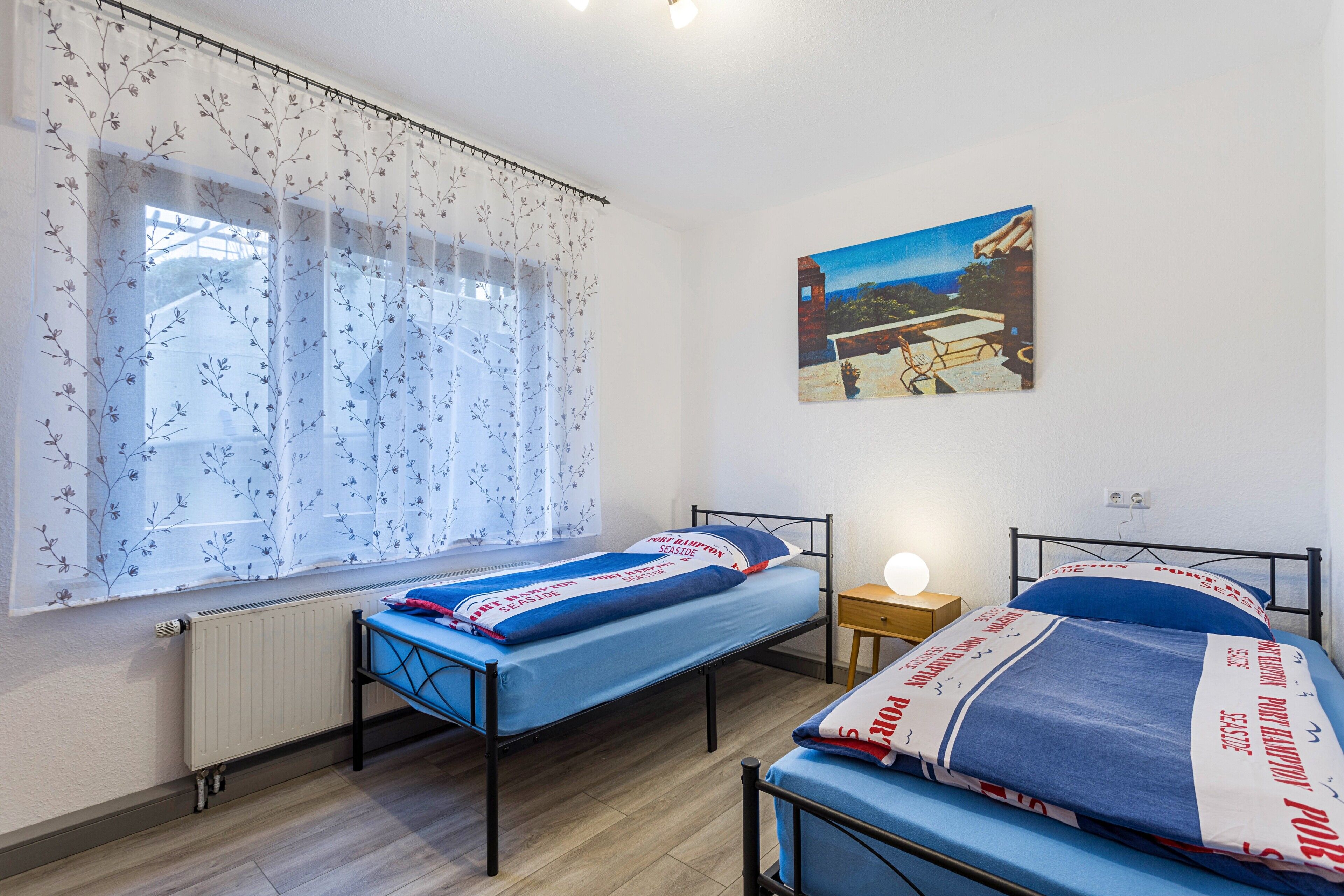 A bedroom with two beds, a window with floral curtains, a nightstand with a lamp, and a painting on the wall.