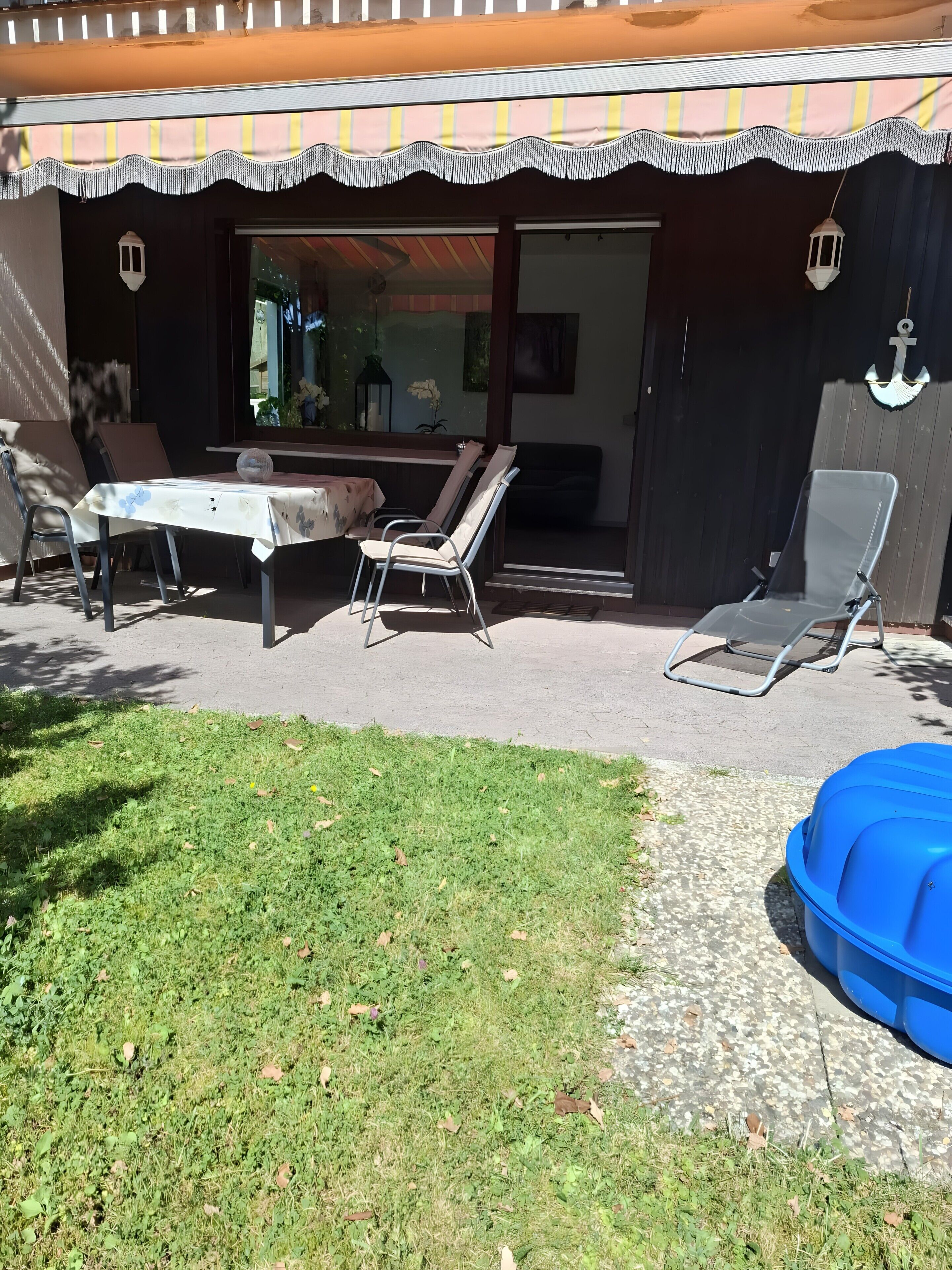 En uteplats med ett bord och stolar, en överbyggd sittplats, en blå uppblåsbar pool och en dekorativ vägg med ett ankarmotiv.