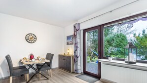 A dining area with a table, chairs, a clock, a cabinet, a window with curtains, and a view of greenery outside. - Apartment 'Astrid' with Private Terrace, Garden, and Wi-Fi (Sundern (Sauerland))