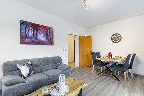 Una sala de estar con un sofá gris, una mesa de centro de madera, una mesa de comedor con sillas, un cuadro de un bosque y un reloj de pared.