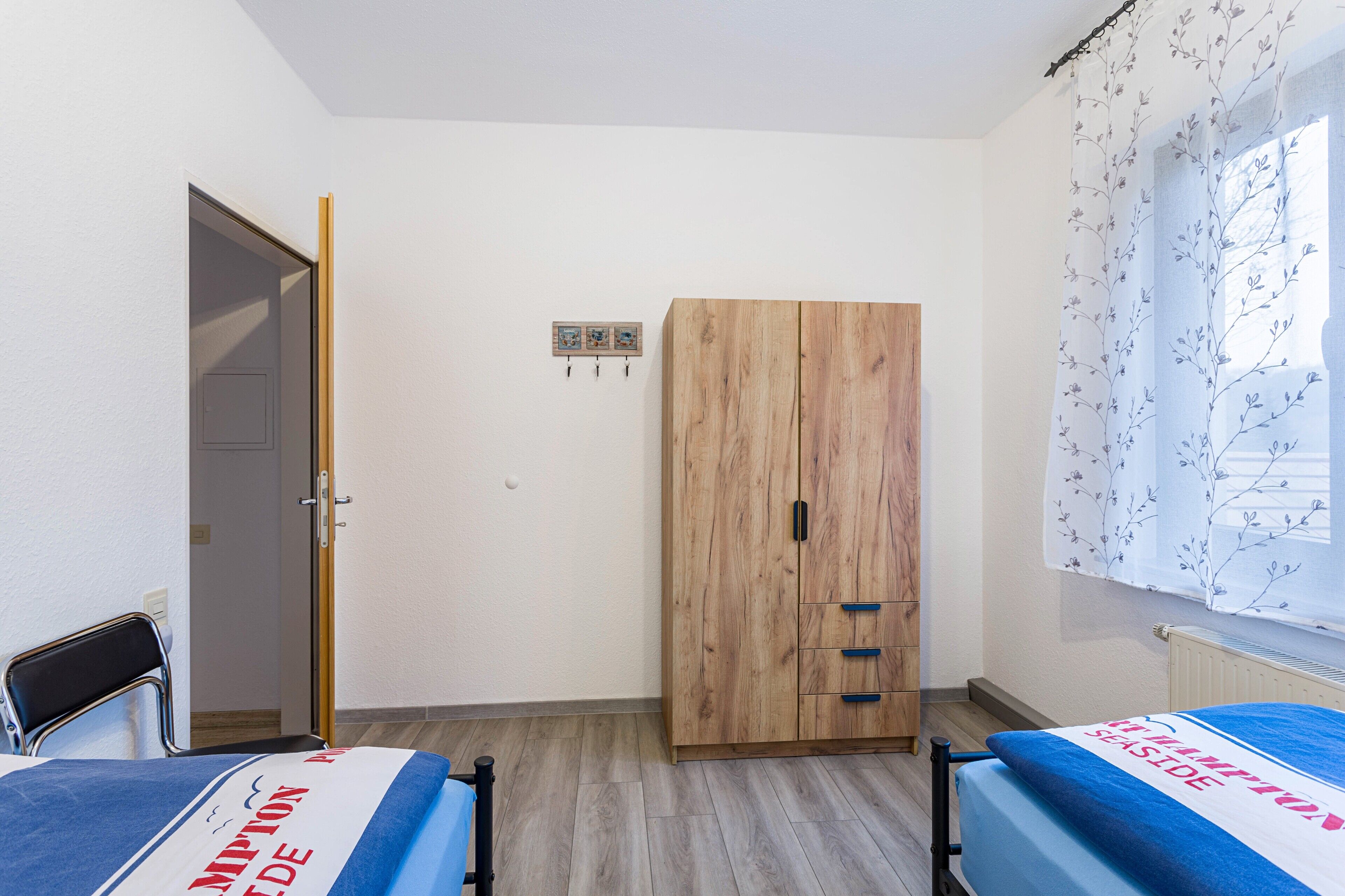 A bedroom with a wooden wardrobe, a bed with a blue bedspread, a chair, and a window with a floral curtain.