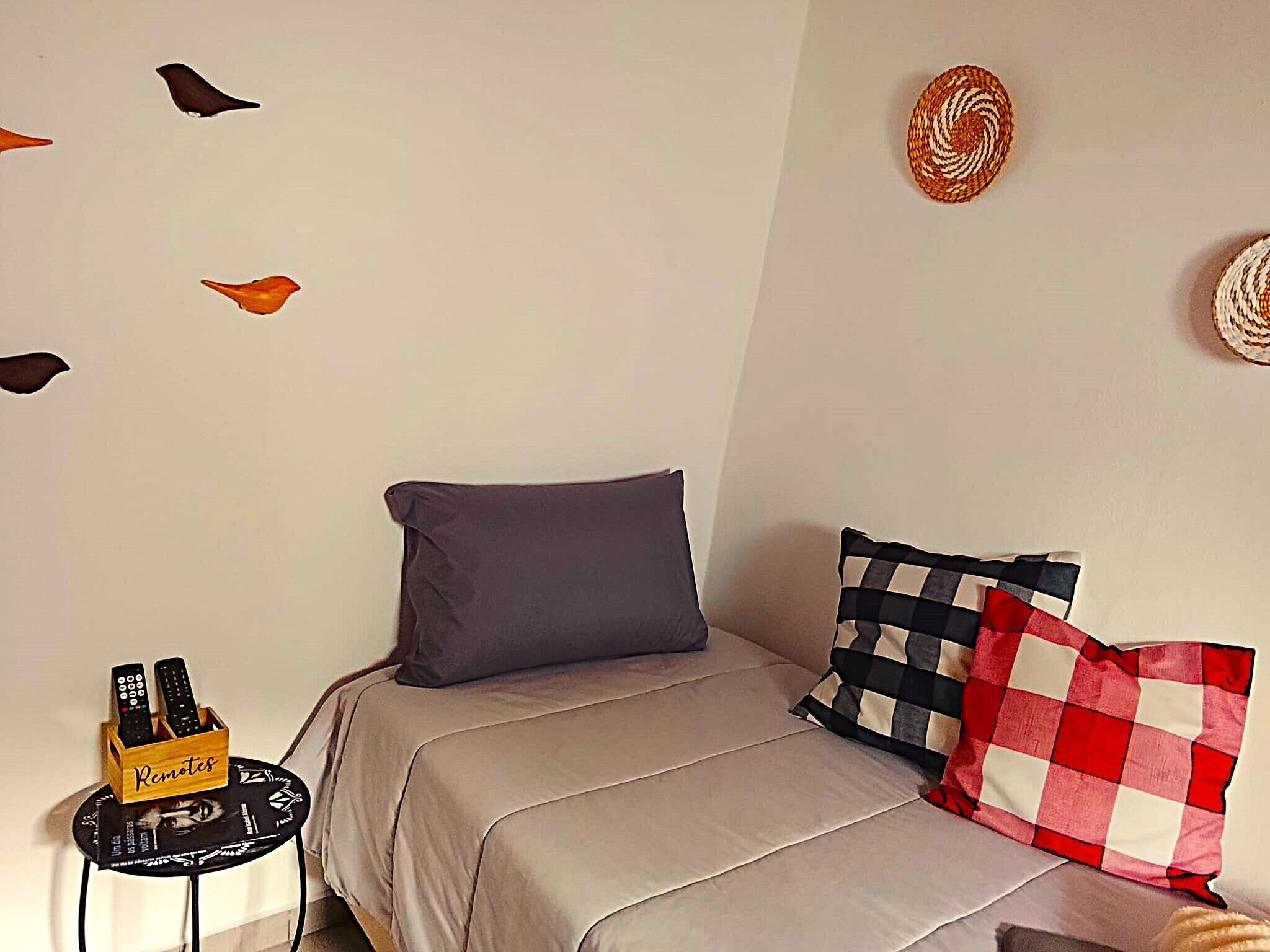 A bedroom with a bed, pillows, a remote control holder, wall decorations, and a small table.
