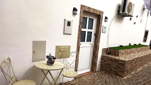 A white wall with a door, a small table with a chair, a wall-mounted air conditioner, and a window with a view of a green area.