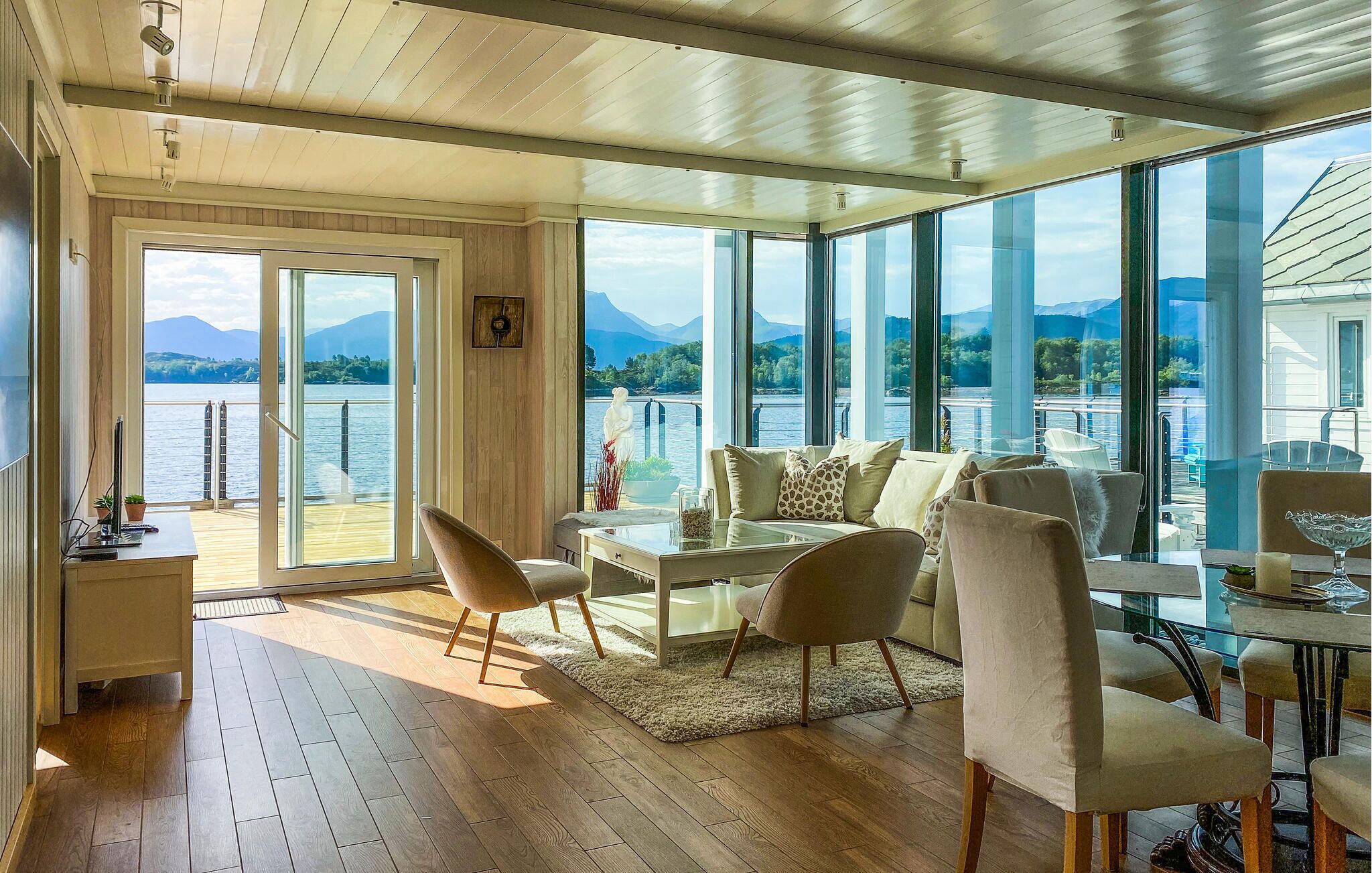 A spacious room with a wooden floor, a sofa set, a dining table, and large windows offering a view of a lake and mountains.