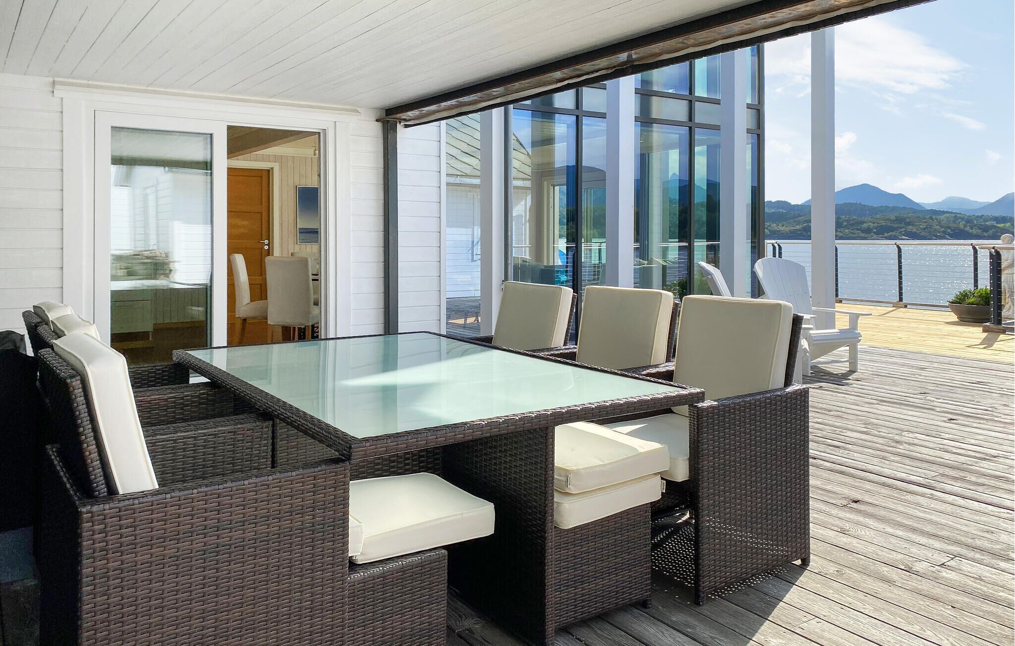 A covered outdoor dining area with a glass table and wicker chairs, overlooking a body of water and mountains.
