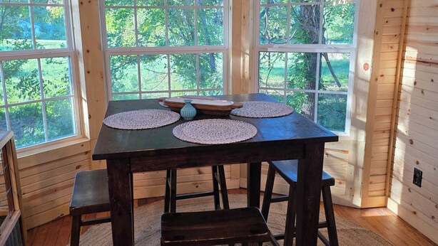 Een houten interieur met een tafel en vier krukken, grote ramen met uitzicht op groen, en een geweven vloerkleed.