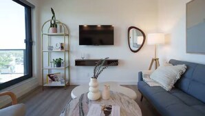 A modern living room with a blue sofa, a round coffee table, a gold shelving unit with plants, a flat-screen TV, and a mirror on the wall. - Stylish Condo W/ Stunning views & Short Walk DT (Calgary)