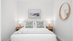 A neatly made bed with white bedding and green patterned pillows, two bedside tables with lamps, a framed picture on the wall, and a round mirror. - Stylish Condo W/ Stunning views & Short Walk DT (Calgary)