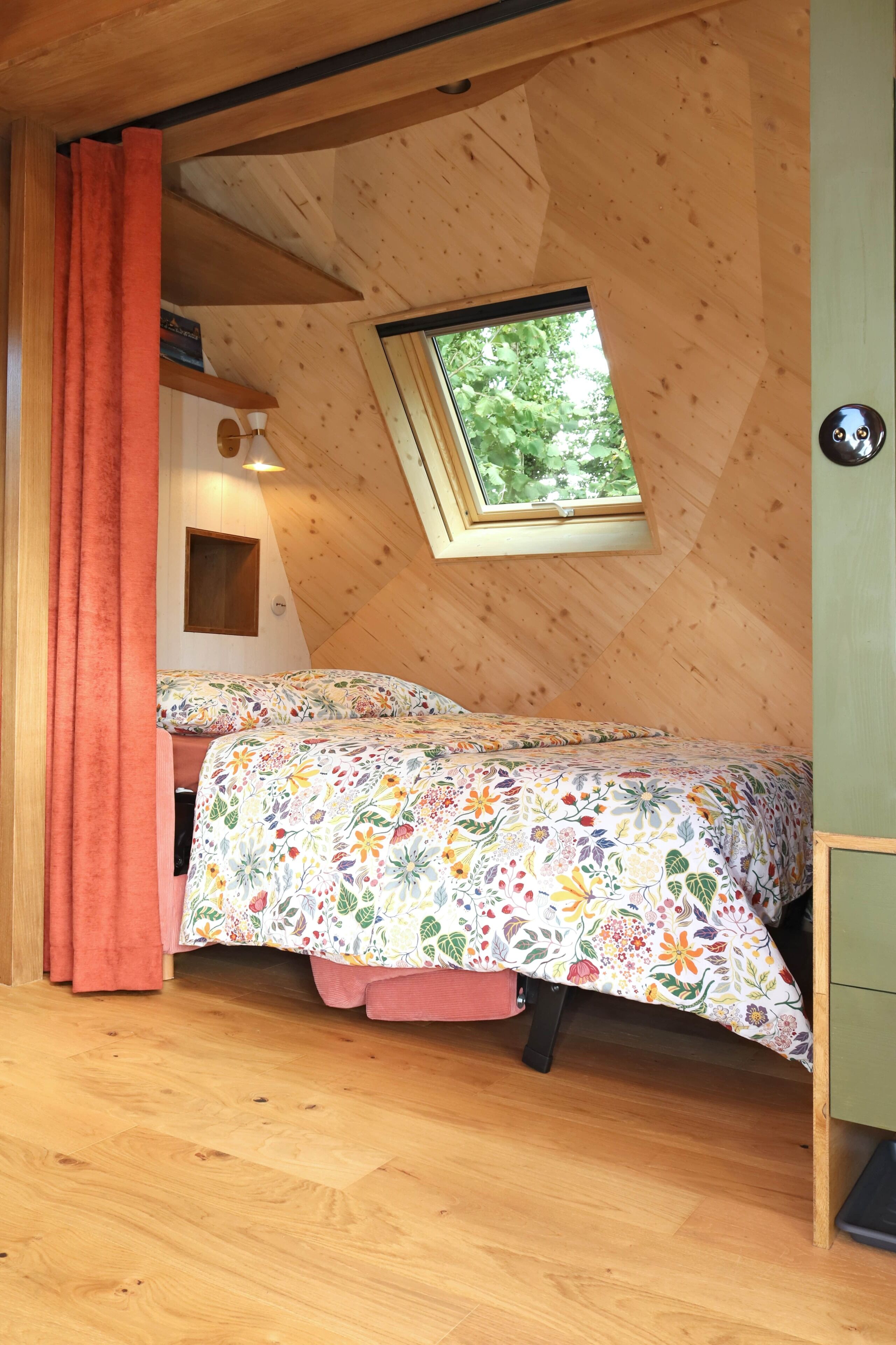 Une chambre confortable avec un plafond en pente, une fenêtre, un lit recouvert d’une literie à motifs floraux et une petite étagère remplie de livres.