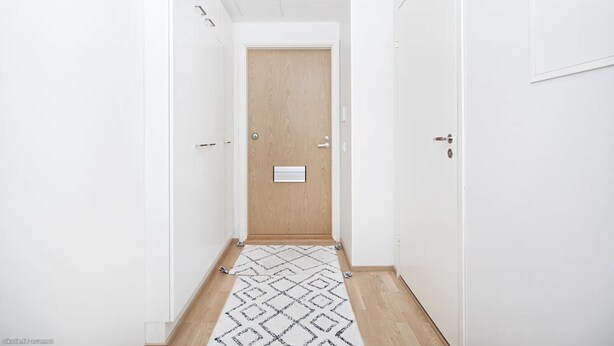 Un pasillo estrecho con una puerta de madera al final, paredes blancas y una alfombra estampada en el suelo.