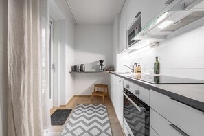 Cozinha compacta com micro-ondas, cafeteira e um pequeno banquinho de madeira.