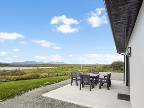 Een terras buiten met een tafel en stoelen, een grindpad en uitzicht op een grasveld en bergen.