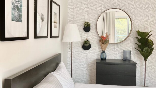Una camera d'albergo moderna con un letto rifatto, un comodino, una lampada, una cassettiera e uno specchio. Alle pareti ci sono quadri incorniciati e un vaso con una pianta sulla cassettiera.