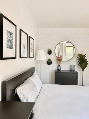 Una habitación de hotel moderna con una cama bien hecha, una mesita de noche, una lámpara, un aparador y un espejo. Hay cuadros enmarcados en la pared y una planta en maceta sobre el aparador.