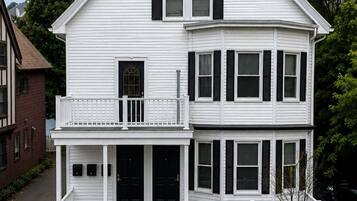 Une maison blanche à deux étages, dotée d’un porche à l’avant, d’une cheminée et d’un petit balcon.