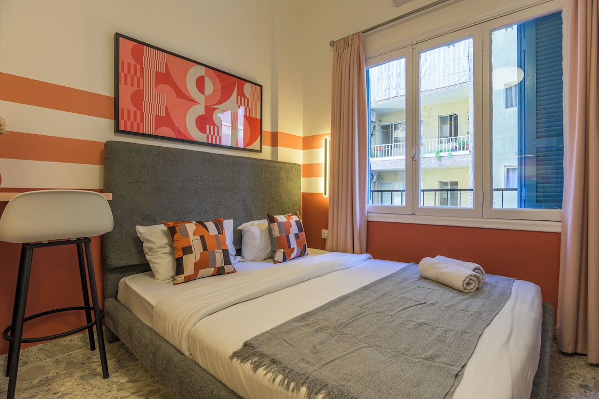 A hotel room with a bed, a bar stool, a window with curtains, and a view of a building.