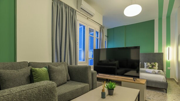 Ein modernes Wohnzimmer mit grauem Sofa, Flachbildfernseher, einem kleinen Tisch mit Pflanze sowie einem Bett, dessen Wände in Grün gestrichen sind und ein Fenster mit blauen Fensterläden hat.
