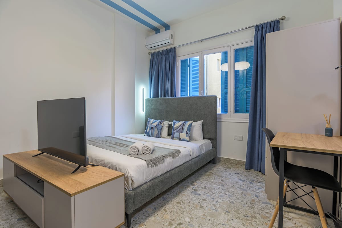 A bedroom with a bed, a TV on a stand, a desk, and a chair.