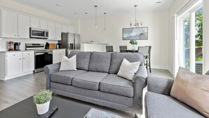 A modern kitchen with white cabinets, a grey sofa, and a dining area with a table and chairs. - Sleeps 6 - 12 min from Fort Jackson - EV Charger & Pet Friendly! (Elgin)