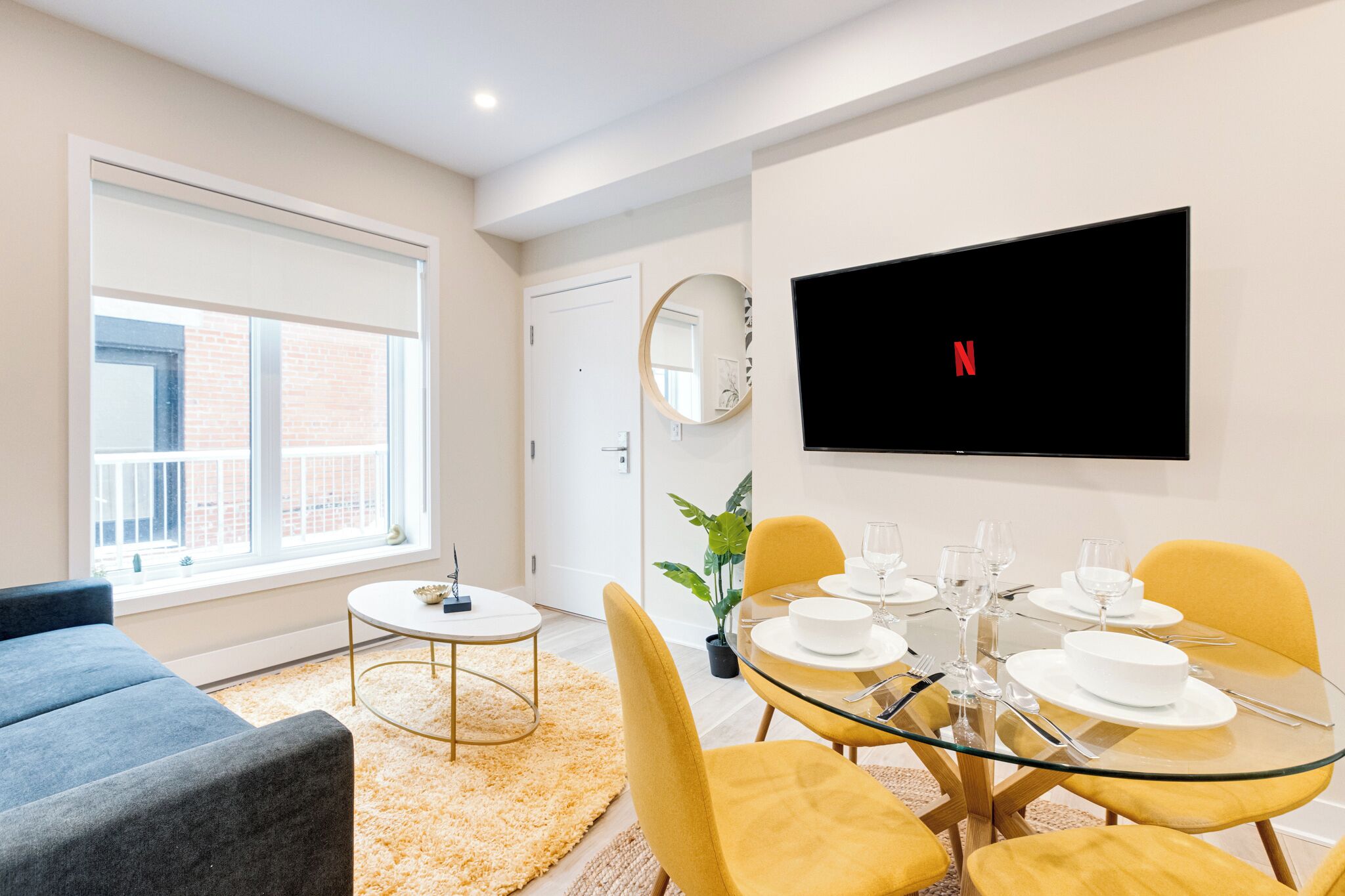 A modern living room with a sofa, a round table set for dining, a TV mounted on the wall, and a large window.