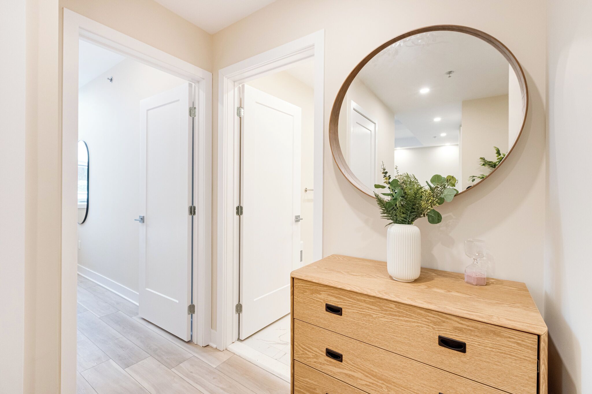 Un couloir avec deux portes blanches, une commode en bois dotée de tiroirs, un miroir rond et une plante en pot.