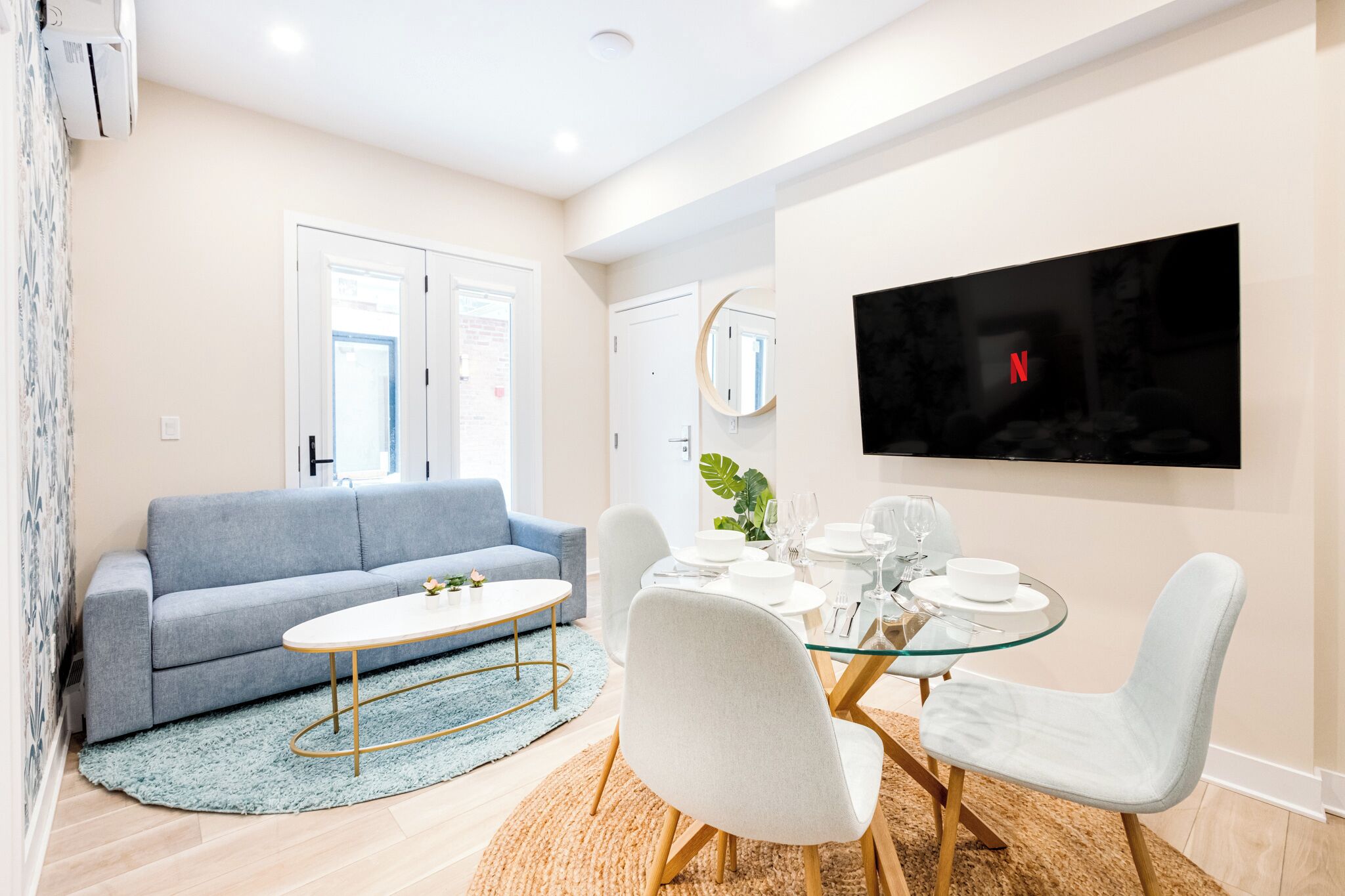 Una sala moderna con un sofá gris, una mesa de comedor de vidrio preparada para cuatro personas y un televisor de pantalla plana montado en la pared.