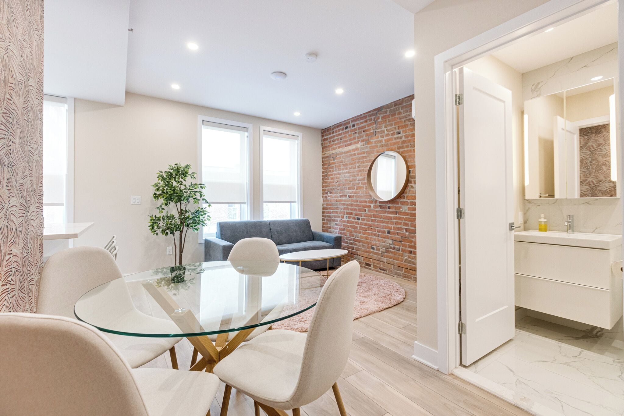 A modern living room with a glass table, white chairs, a grey sofa, and a brick accent wall.