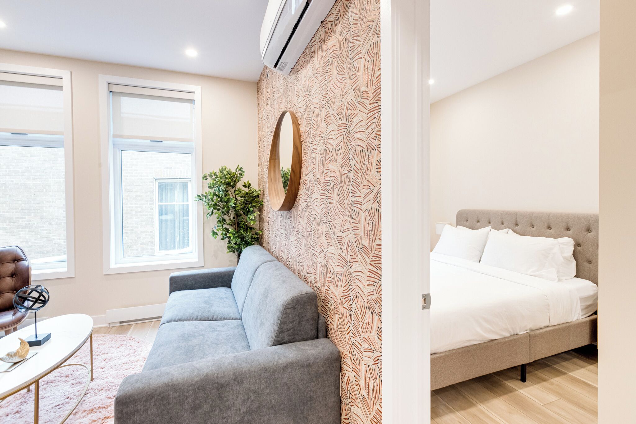 A modern bedroom with a grey sofa, a round mirror, and a bed with white bedding.