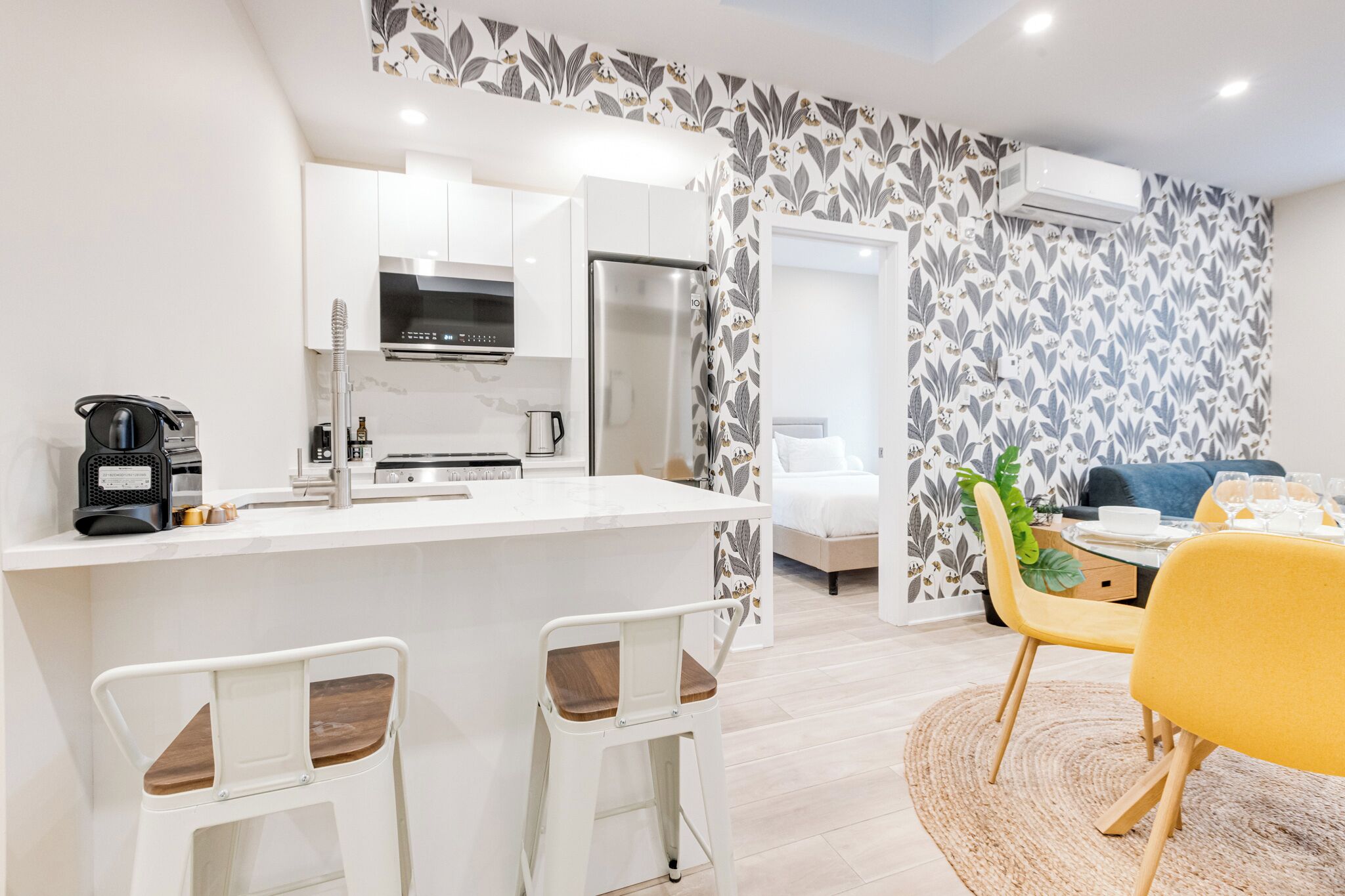 A modern kitchen with white cabinets, a stainless steel refrigerator, and a coffee machine. There is a dining area with a yellow chair and a table.